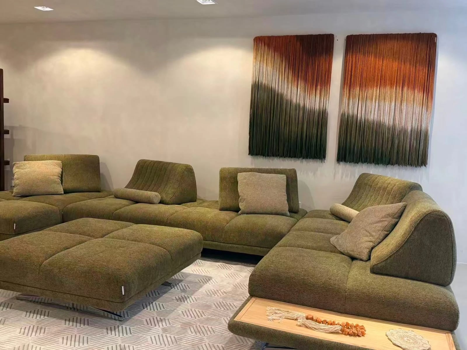 Sofá modular em tecido verde-oliva diante de dois painéis de arte têxtil, no showroom.