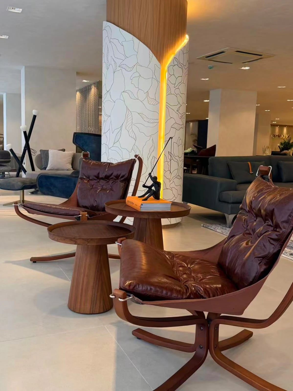 Duas poltronas de couro marrom com estrutura curva em madeira, ao lado de mesas laterais redondas, no showroom.